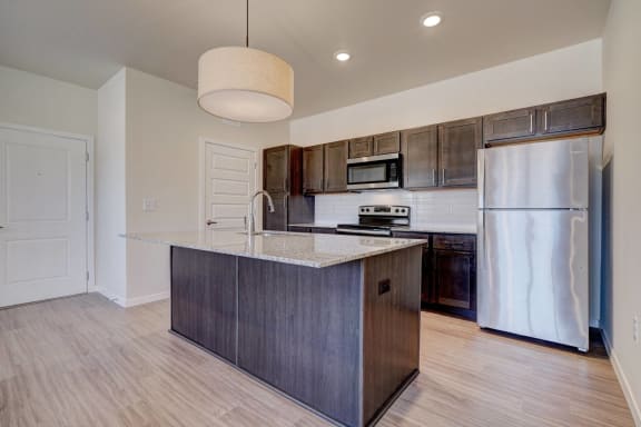 A large, modern kitchen with stainless steel appliances, granite countertops with lots of room, a kitchen island, and large dark wooden cabinets at the Encore Apartment Homes. 
