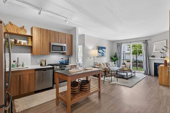 Open apartment kitchen and living room with stainless steel appliances, island, sofa, and patio door.