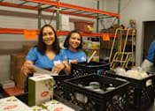 Thumbnail 17 of 21 - Two women in blue shirts are working in a warehouse with boxes and crates.