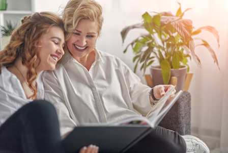 Two women are sitting on a couch, one is reading a book and the other is laughing.
