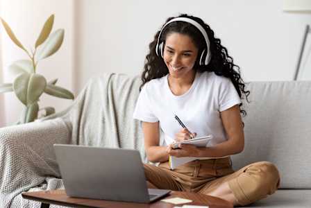 A woman is sitting on a couch with a laptop and headphones on, writing in a notebook.