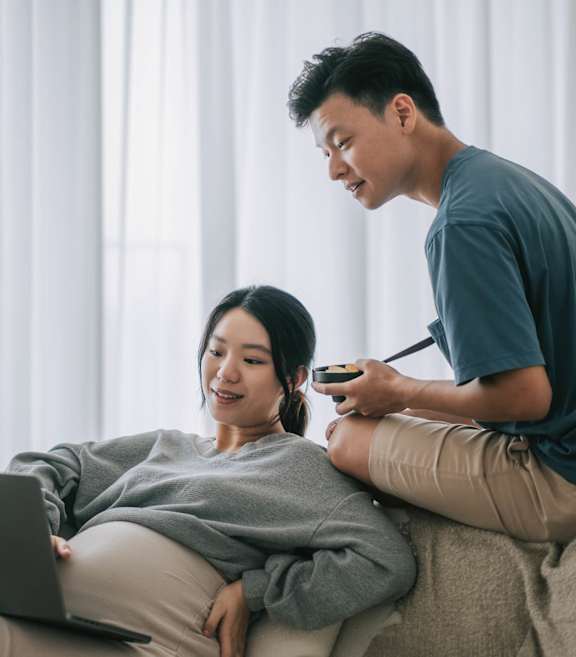 A man is sitting on a couch with a woman, both looking at a laptop screen.