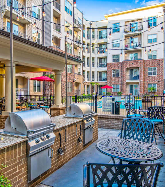 A patio with a grill and chairs is surrounded by a brick wall and buildings.