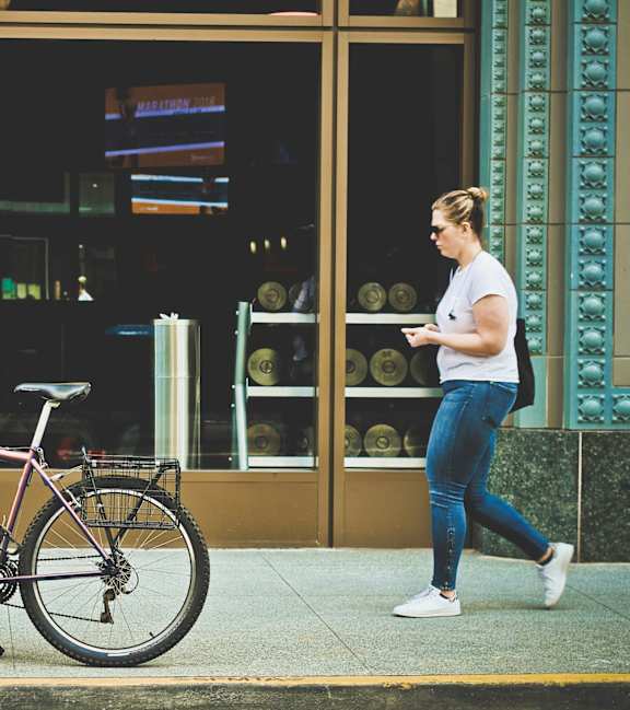 A woman walking past a bicycle parked on the sidewalk.