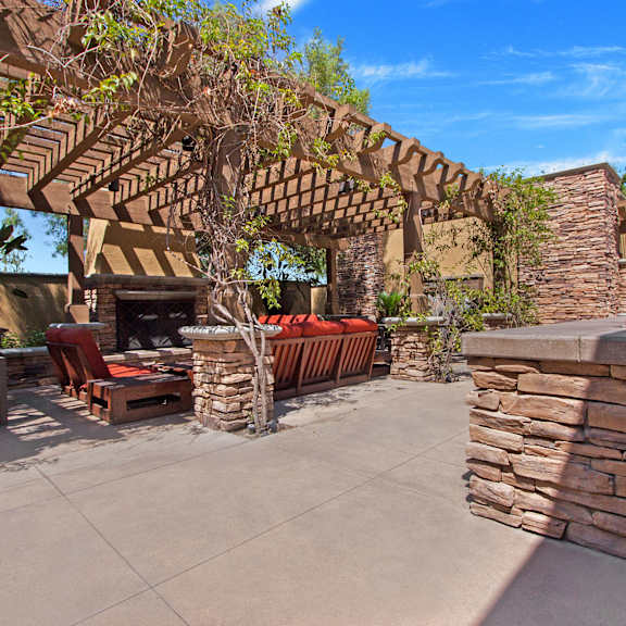 A patio with a stone fireplace, a grilling station, table and chairs, plush orange outdoor chairs and an orange couch, and a pergola with vines climbing up it at Vesada Apartment Homes. 
