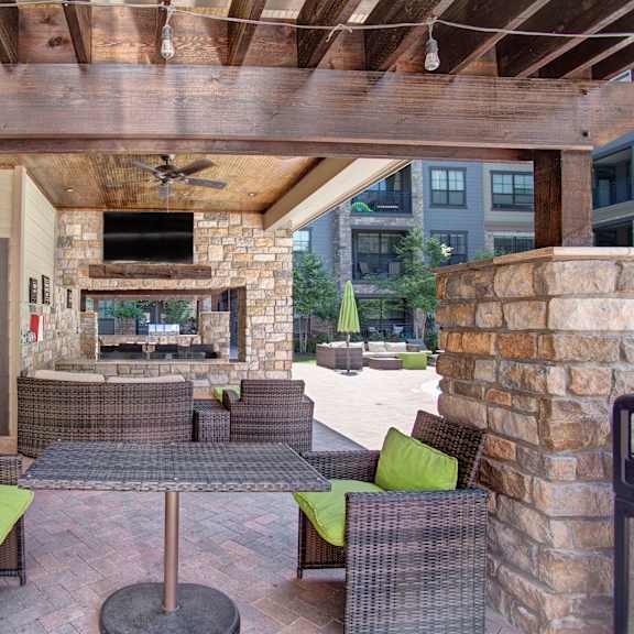 The outdoor patio with a TV, green plush chairs, a tan couch, a fireplace, a coffee table, and a covering at Enclave at Brookside Apartment Homes. 