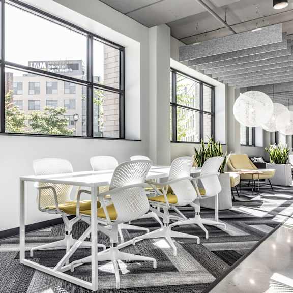 The community sitting area with a long white table surrounded by white chairs with unique white lighting above with plants and plush chairs in the background at Vim + Vigor Lofts. 