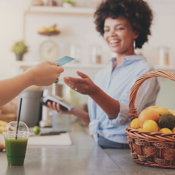 A person is paying with a card at a counter with a basket of fruit and a green drink.