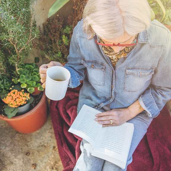an older woman sitting on a blanket reading a book and holding a cup of coffee at The Carillon, Greensboro, 27455