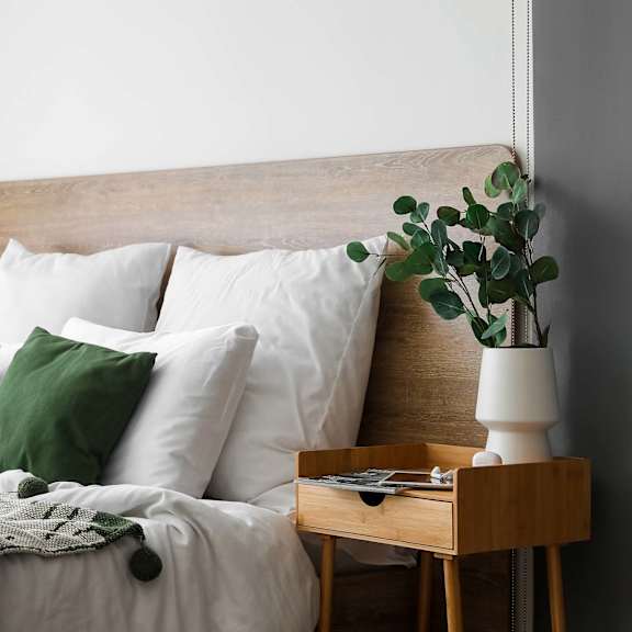 A bed with white pillows and a green throw is next to a wooden nightstand with a plant on it.