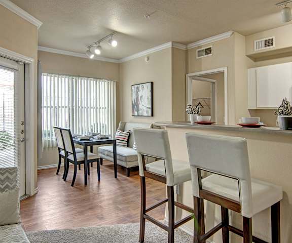 A cozy kitchen with white appliances, light counter with ample space, spacious wooden cabinets, and a dining room with a dark table, dark chairs with white cushions, and a white bench at Mozaic at Steele Park Apartments. 