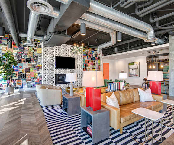 Roosevelt Row Resident Lounge The amazing, industrial resident lounge with a fireplace with a TV above it, tan leather couches, decorations, large windows with curtains, colorful wall art, white plush chairs, and coffee tables at Roosevelt Row Apartment Homes.