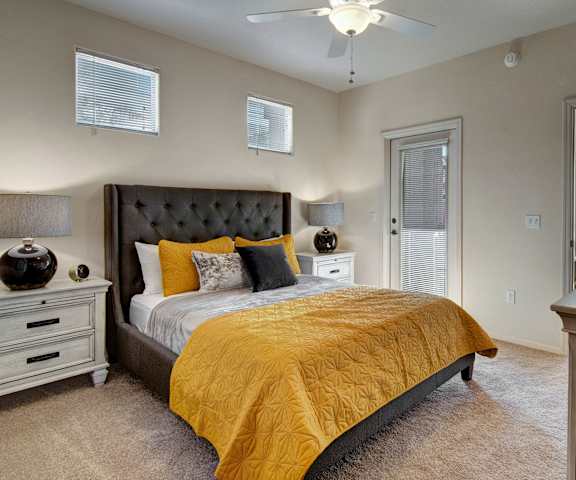 A spacious bedroom with a queen-size bed with a silver, black, and yellow bedspread, a dresser, wall art, lamps, decorations, window, nightstands, and doors to the balcony at Sonoma Landing Apartment Homes. 