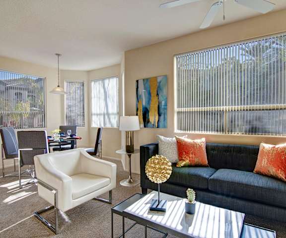 The spacious living room with a denim couch, orange and white throw pillows, a white leather chair, coffee table, overhead lighting with a ceiling fan, decorations, wall art, windows, and view of the dining room with a table, chairs, and place settings for four at Sonoma Landing Apartment Homes. 