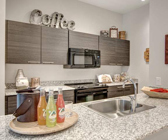 A spacious, modern kitchen with black appliances, marble countertops with lots of room, kitchen utensils, a drink stand with fruit sodas, and gray wooden cabinets with decorations at the Trend at 51 Apartment Homes. 
