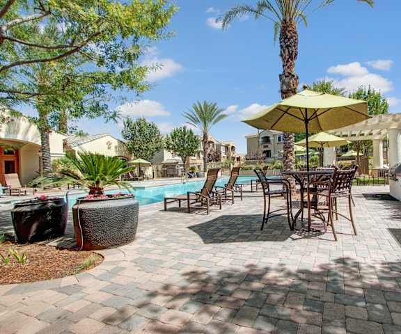 The outdoor patio with tables, chairs, umbrellas, grills, with the pool and pool chairs in the background surrounded by trees in Rancho Belago Apartments. 