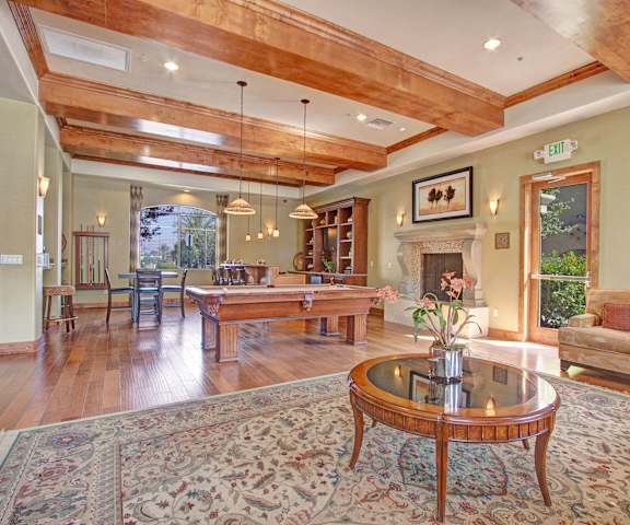 A large, well-lit resident lounge with wooden accents, windows, a door to the outside, decorations, a plush couch, bookshelves, tables, chairs, and a pool table at Rancho Belago Apartments. 