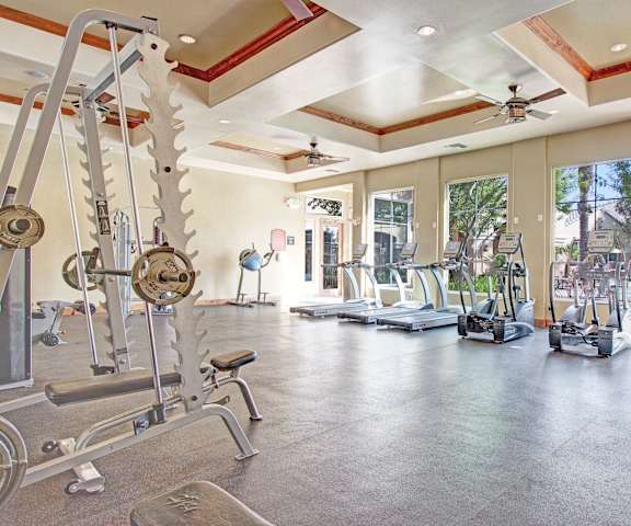A gym with cardio machines, windows with a view of the pool area, TVs, and weights at Rancho Belago Apartments. 