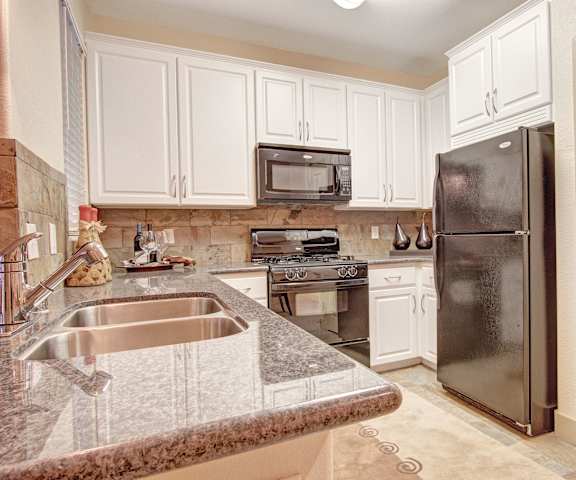 A large kitchen with stainless steel appliances, spacious marble countertops, and large, white wooden cabinets at Rancho Belago Apartments. 