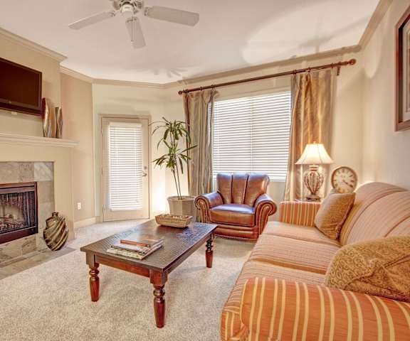 The living room with a plush couch, a leather chair, windows, door to the balcony, coffee table, decorations, a TV, and view of the dining room and the kitchen at Rancho Belago Apartments. 