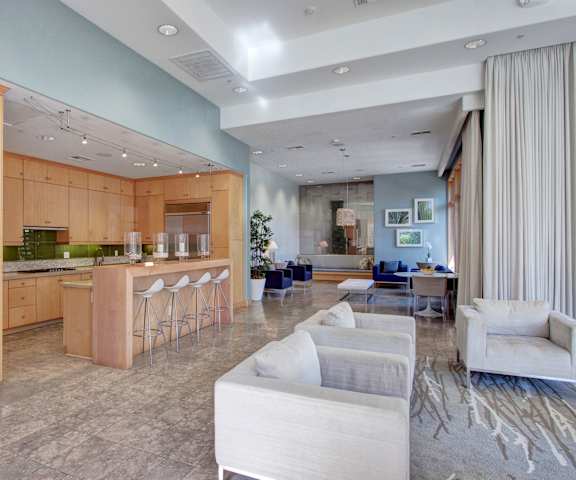 The amazing resident lounge with artwork, plants, couches, decorations, large windows with curtains, plush chairs, coffee tables, and a resident kitchen with barstools, stainless steel appliances, and many large light wooden cabinets at Vesada Apartment Homes. 