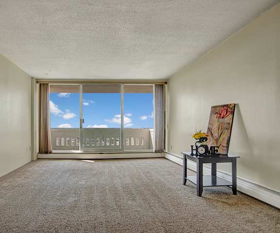 An empty living room with carpeting, a coffee table with a "Home" sign, flowers, and a painting, and a door to the balcony at Marquis Towers Apartment. 