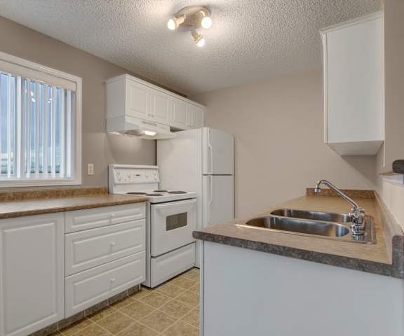 A cozy kitchen with white appliances, kitchen utensils, overhead lighting, light countertops with lots of room, a small window, and white, large wooden cabinets at the Pointe at Applewood Apartment Homes. 
