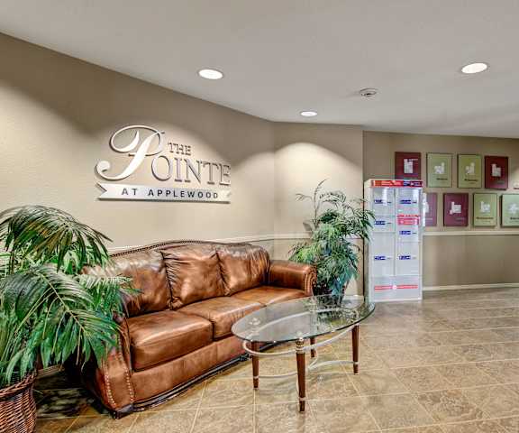 The lobby with a brown leather couch, two palm plants in pots, a glass coffee table, a metal sign on the beige wall, wall artwork, and mailboxes at the Pointe at Applewood Apartment Homes. 
