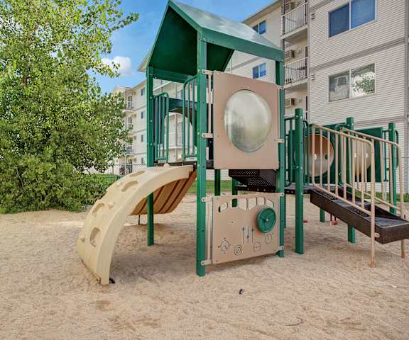The green and tan playground with sand and a tree in the background at Villagio Apartment Homes. 