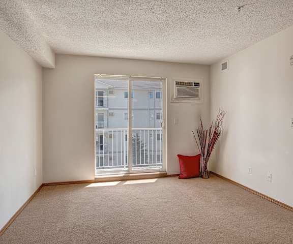 A large, empty living room with carpeting, a red pillow, a plant, and a door to the balcony at Villagio Apartment Homes. 