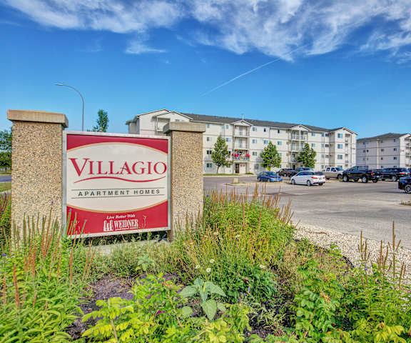 The red and white exterior sign with trees and surrounded by greenery with the buildings in the background of Villagio Apartment Homes. 