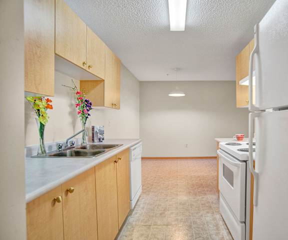 A cozy kitchen with white appliances, overhead lighting, light counter, flowers, and large wooden cabinets at Wellington Manor Apartment Homes. 