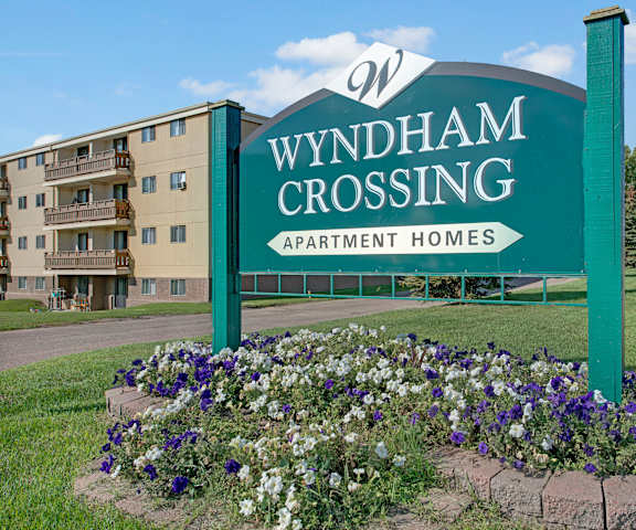 The exterior sign with trees and surrounded by greenery with blue and white flowers and the buildings in the background of Wyndham Crossing Apartment Homes. 
