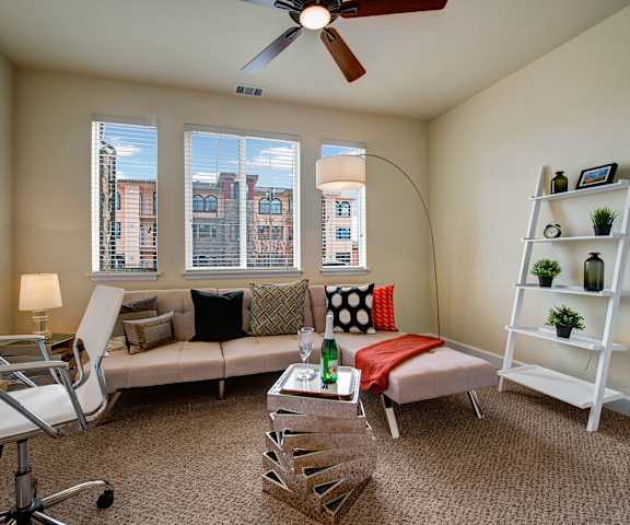 An office with a desk, chair, couch with pillows, windows, shelves, coffee table, and decorations at La Bella Vita Apartments.