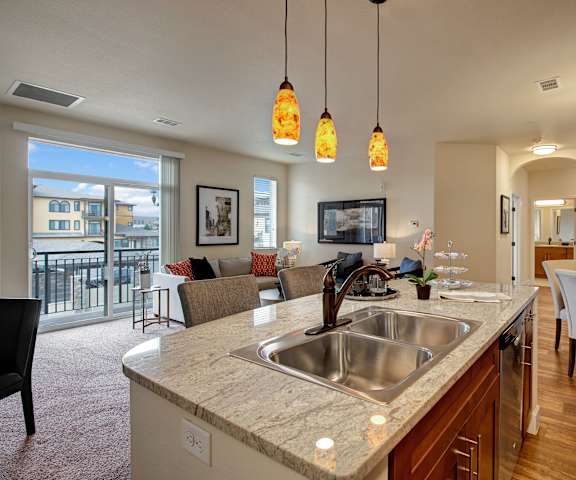 A marble kitchen with a view of the dining room with a table and chairs, a living room with a couch and chairs, and the doors to the balcony at La Bella Vita Apartments. 