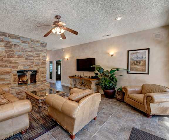 The amazing resident lounge with a fireplace, tan leather couches, decorations, windows, tan leather chairs, decorative plants, overhead lighting and a ceiling fan and coffee tables at Windtree Apartment Homes. 