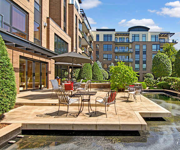 The Mist Deck The outdoor resident patio with tables, umbrellas, and chairs with a water feature and green space with trees and bushes at the Mist at Lake Minnetonka Apartment Homes.
