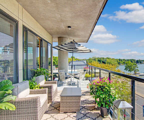 The Mist Balcony A beautiful balcony overlooking Lake Minnetonka with plush white chairs, a coffee table, an umbrella, and a white table and chairs with potted flowers growing at the Mist at Lake Minnetonka Apartment Homes.
