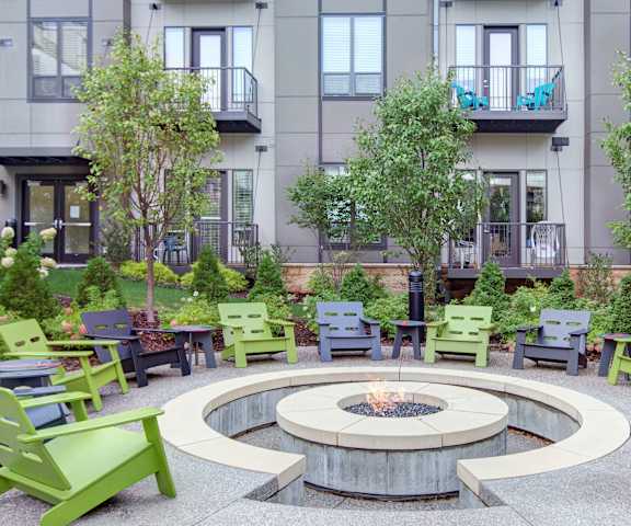 A circular firepit surrounded by black and green with the green space, trees, and the gray apartment building at the Residence at the COR Apartment Homes. 