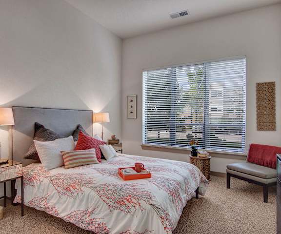 A spacious bedroom with a king-sized bed with a white and floral pattern, two nightstands, two lamps, a plush gray chair with a red throw blanket, a dresser, and a large window at the Residence at the COR Apartment Homes. 