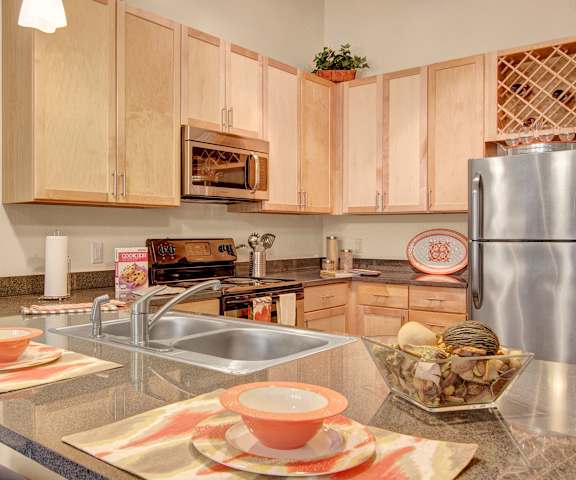 A large/cozy kitchen with stainless steel appliances, bar counter with barstools with two orange place settings,, kitchen utensils, marble countertops with lots of room, and large wooden cabinets at Residence at the COR Apartment Homes. 