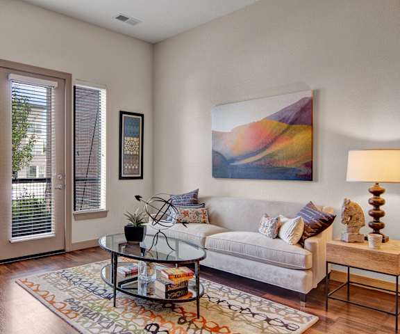 The spacious living room with a gray couch, throw pillows, large windows, a horse head figure, wall art, a plush orange chair, coffee table, decorations, large windows, a TV, and glass doors to the balcony at Residence at the COR Apartment Homes. 