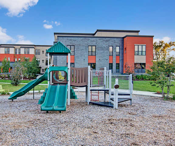Playground A brown and green playground in front of the gray and orange apartment building at Siena Apartment Homes.
