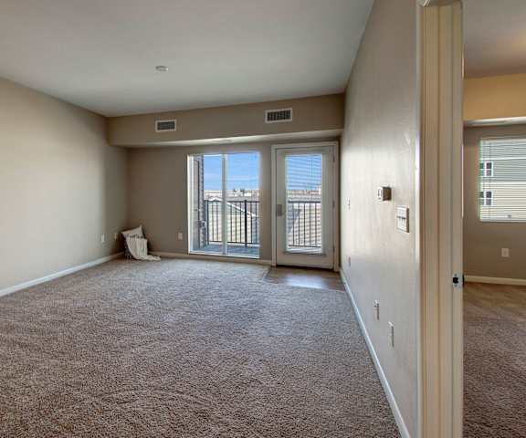 An empty living room with carpeting and a door to the balcony at Dakota Commons Apartments. 