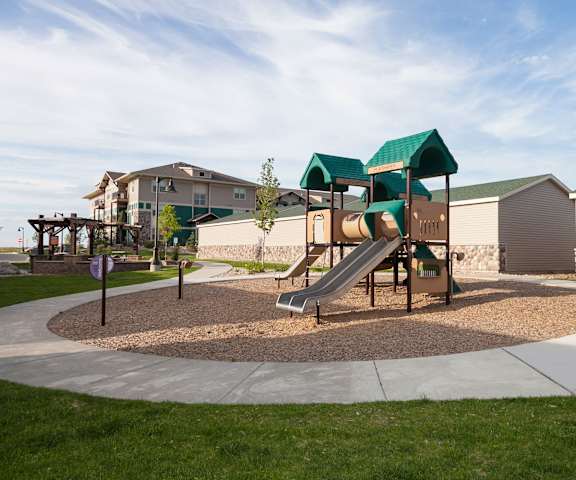 Prairie Pines at the Ridge Playground A green and light brown playground with slides surrounded by green space at a park at Prairie Pine at the Ridge Apartment Homes.