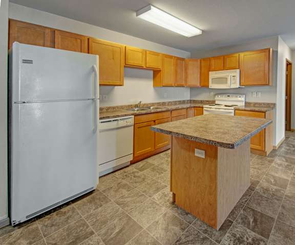 Kitchen at Roosevelt East Apartment Homes A spacious kitchen with white appliances, kitchen utensils, overhead lighting, light countertops with lots of room, a bar counter, and large wooden cabinets at Roosevelt East Apartment Homes.