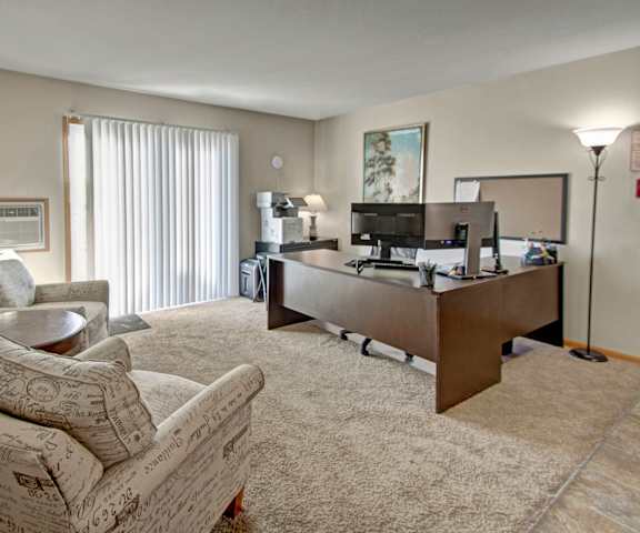 Leasing Office at Roosevelt East Apartment Homes A modern office with a desk, desktop computer, plush chairs, shelves, coffee table, wall art, decorations, and sliding glass door with light streaming in at Roosevelt East Apartment Homes.