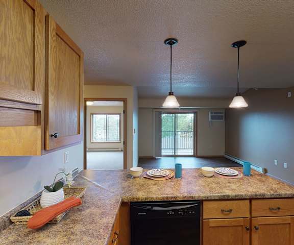 A view of the empty carpeted living room with overhead lighting, doors to the balcony, two empty bedrooms, and the large kitchen with black appliances, marble countertops with two place settings, and spacious wooden cabinets at Williston Gardens Apartment Homes. 