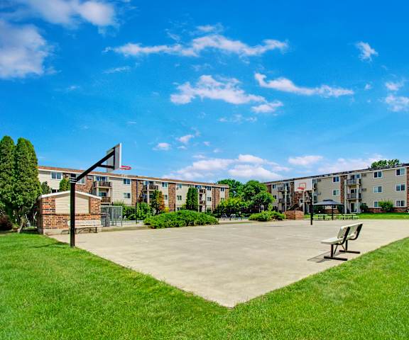 Windscape Basketball Court The courtyard with a basketball court, benches, a grassy area with some trees and shrubbery, and the apartments in the background with blue sky above at Windscape Apartment Homes.