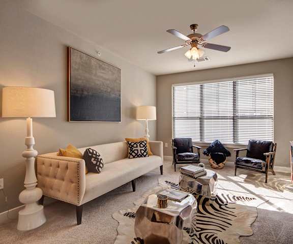 The spacious living room with a tan couch, zebra print rug, throw pillows, large windows, a ceiling fan, wall art, two black plush chairs, coffee table, decorations, large windows, and a TV at the Enclave at Brookside Apartment Homes. 